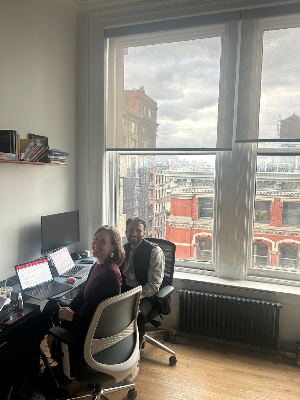 A picture of Kate and Tim in JFI offices in Manhattan, both sitting with laptops at a small desk and turning to smile at the camera. The desk is next to a large window with some interesting New York architecture visible outside, and a somewhat overcast sky. 