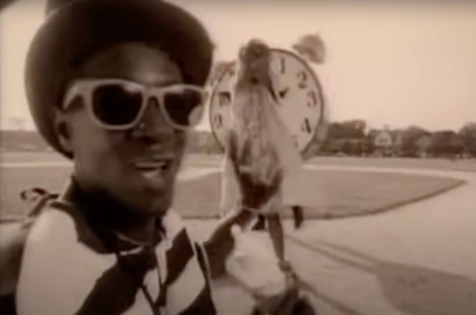 Still from the video of "Can't do Nuttin For Ya, Man" by Pubilc Enemy. Shows Flavor Flav in a stripey top wearing big sungasses. In the background is some kind of person walking past a giant clock. the whole scene is separa-toned.